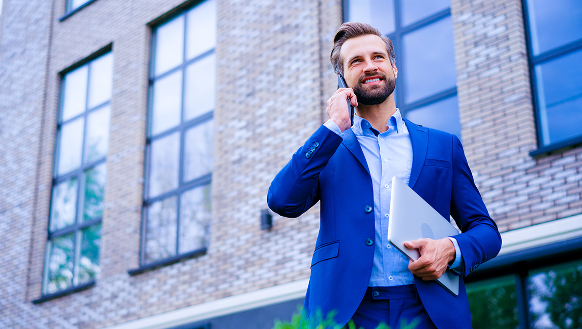 Businessman walking outside office building carrying laptop while talking on his mobile phone