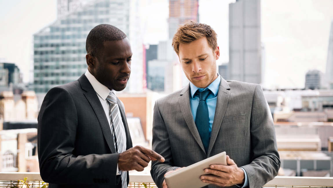 Two male colleagues standing on terrace looking at a tablet