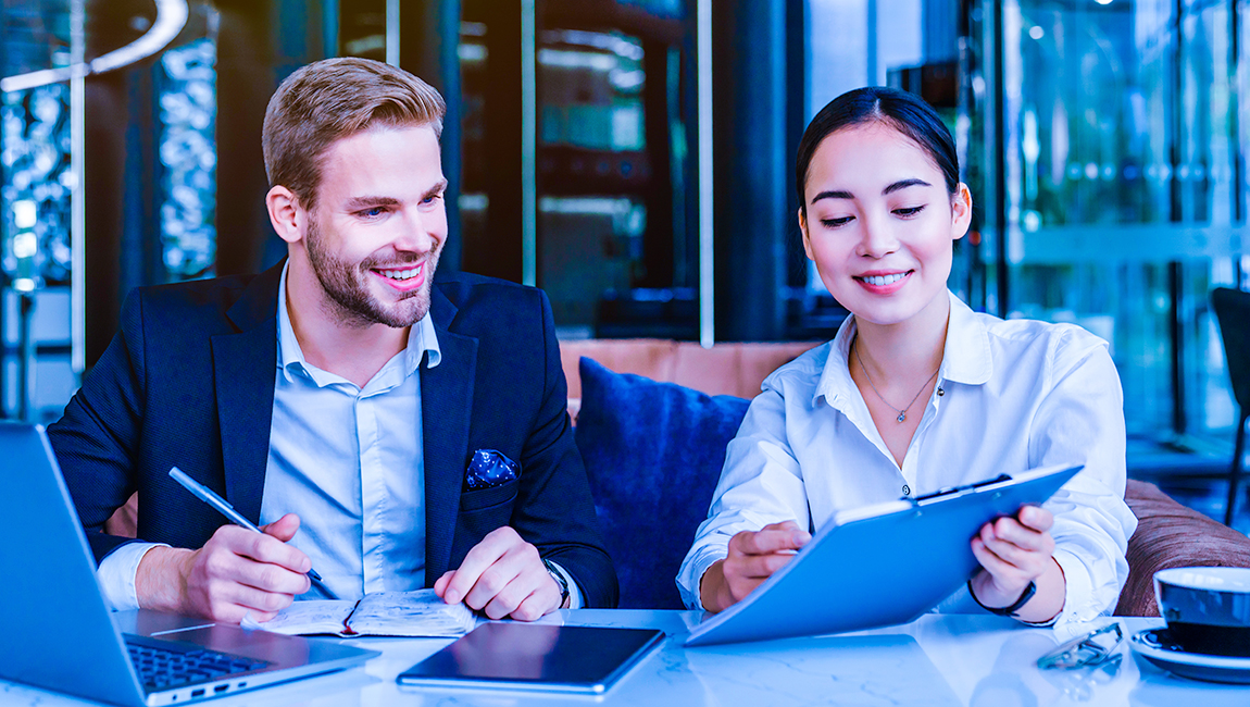 Two colleagues seated next to each other at table reviewing clipboard